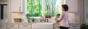 A person stands at a kitchen sink preparing food, with potted plants and kitchen items on the counter, and a large window showing trees outside.