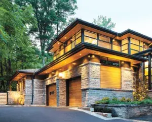 Modern two-story house with large windows, a flat roof, stone and wood exterior, set among tall trees with a driveway in front.