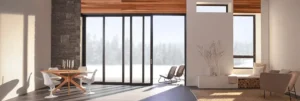 Modern living room and dining area with large floor-to-ceiling glass doors, minimalist furniture, stone accent wall, and wood ceiling detail. Natural light fills the space.