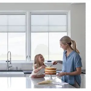 A woman and a young girl decorate a layered cake together in a bright, modern kitchen with large windows in the background.