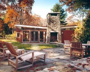 A modern house with large glass doors, a stone outdoor fireplace, and wooden patio furniture set on a stone terrace surrounded by autumn trees.