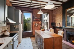 A rustic kitchen with exposed brick walls, wooden beams, two large windows, a central wooden island, modern appliances, and a fireplace with a mantel.