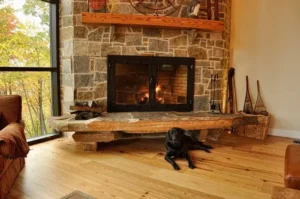 A black dog lies on a wooden floor in front of a stone fireplace with a fire burning inside, surrounded by rustic decor in a cozy living room.