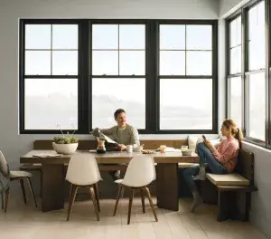 Two people sit at a dining table by large windows; one pours a drink while the other reads, with food and dishes on the table.
