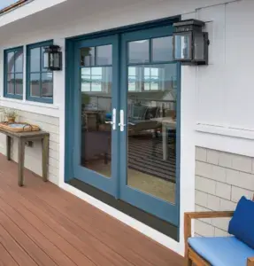 Blue-framed glass double doors on a house open to a deck with wood flooring; outdoor wall lanterns and a small table are nearby.