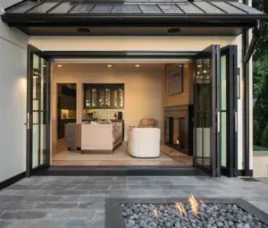 Open glass doors reveal a modern living room with a sofa and armchair; in the foreground, there is an outdoor fire pit with flames and black stones on a stone patio.