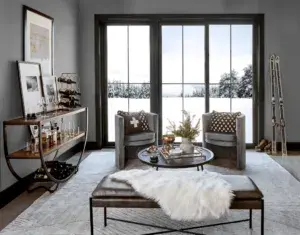 Modern living room with two armchairs, a bench with a fur throw, a round coffee table, a bar cart, large windows, and snow visible outside.