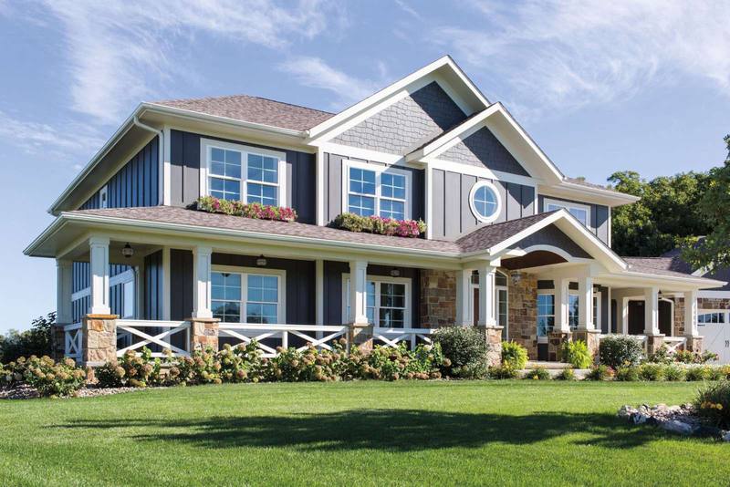 Two-story suburban house with gray siding, white trim, and stone accents, surrounded by a manicured lawn, shrubs, and flower beds under a blue sky.