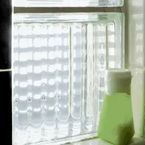 A green bottle with a white cap sits on a windowsill next to a textured glass window letting in light.